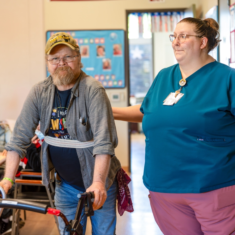 man walking with assistance of walker and woman in scrubs